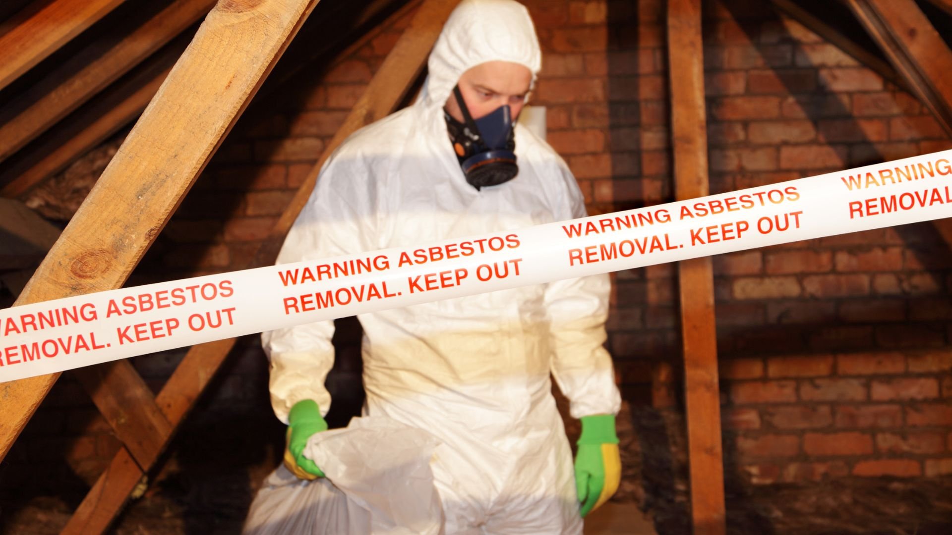 Worker in protective gear removing asbestos from attic with warning tape